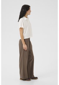 Wit polo-shirt met korte mouwen en kraag, gecombineerd met bruin broek met wijde pijpen en een trekkoord in de taille. Het schoeisel heeft een stevige zool.