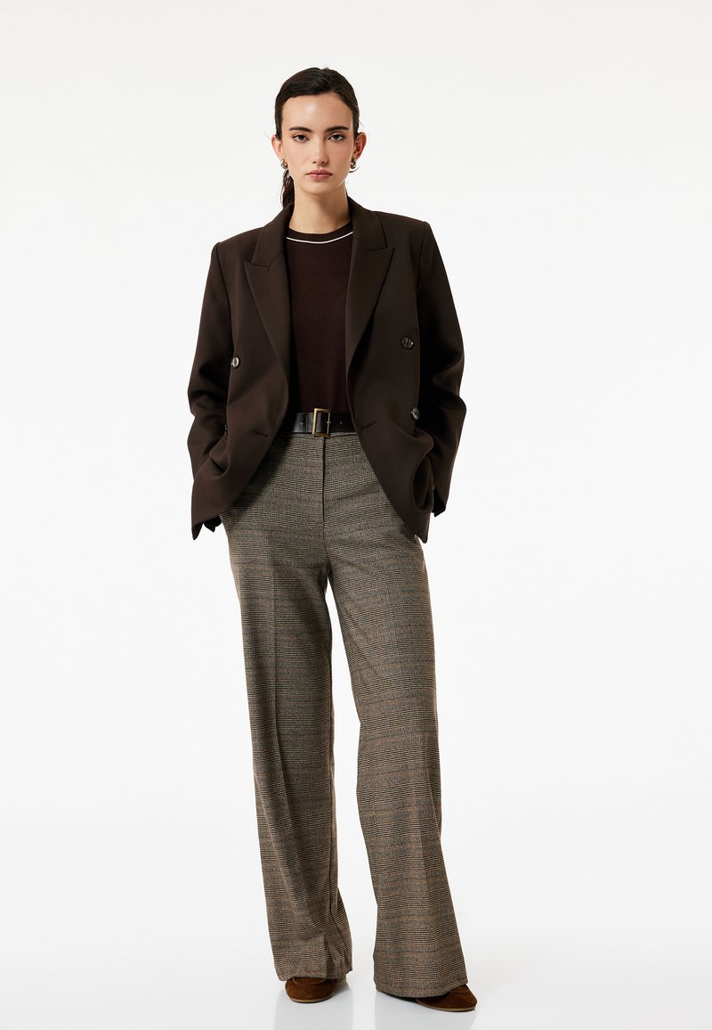 Bruine dubbelgeknoopte blazer over een donkere trui, gecombineerd met een hoge taille schotse broek. De outfit heeft een ruime pijp en een riem.
