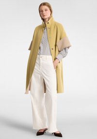 Cappotto giallo in misto lana con colletto strutturato e maniche beige, abbinato a una maglia a righe e a pantaloni bianchi a gamba larga, con scarpe scure.