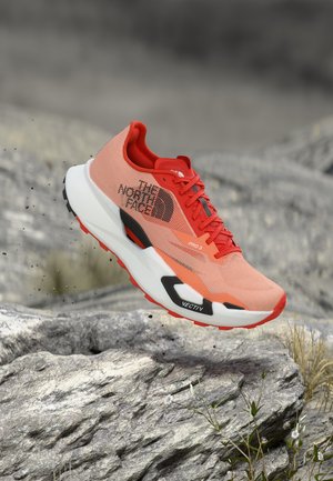 Πορτοκαλί και κόκκινο παπούτσι για τρέξιμο σε μονοπάτια με το λογότυπο "The North Face", στον αέρα πάνω από πετρώδες έδαφος με μικρά φυτά, υπογραμμίζοντας την ανθεκτική χρήση σε εξωτερικούς χώρους.