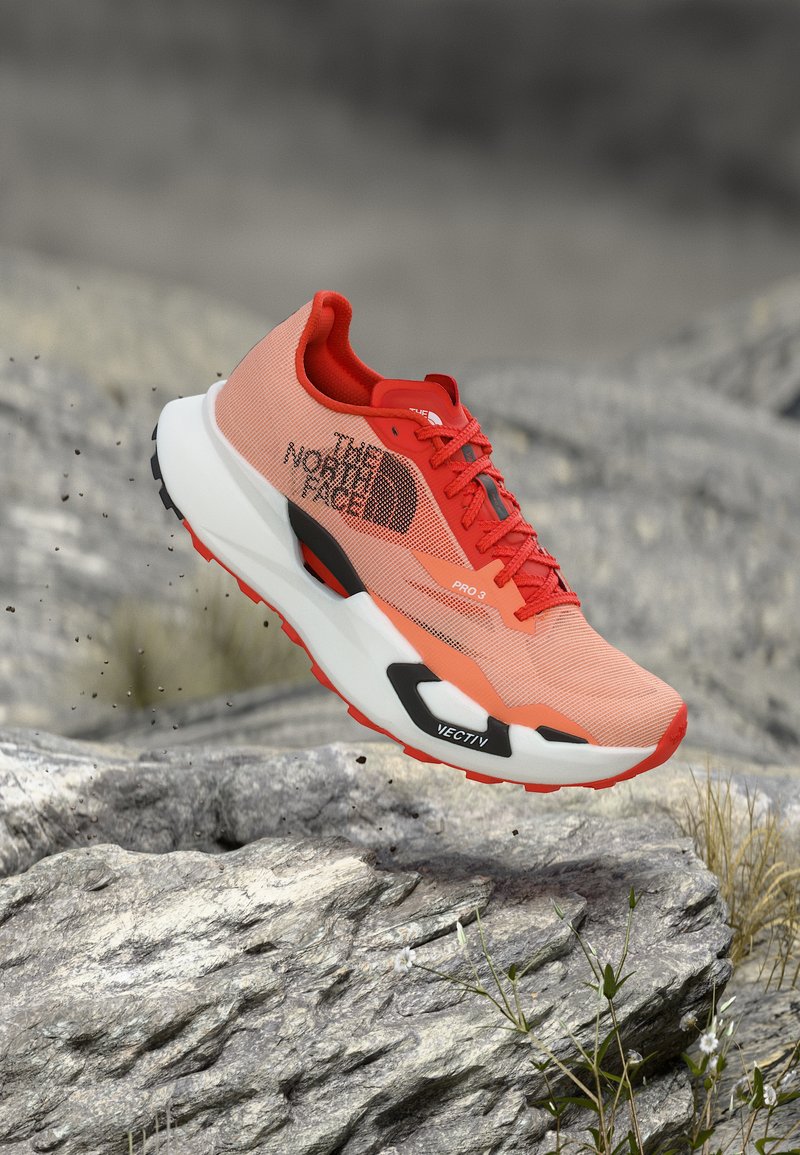 Πορτοκαλί και κόκκινο παπούτσι για τρέξιμο σε μονοπάτια με το λογότυπο "The North Face", στον αέρα πάνω από πετρώδες έδαφος με μικρά φυτά, υπογραμμίζοντας την ανθεκτική χρήση σε εξωτερικούς χώρους.