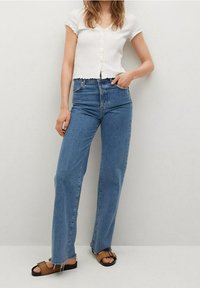 Woman wearing white short-sleeve buttoned top, high-waisted blue jeans, and brown sandals, standing with one hand in pocket against plain background.