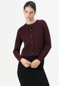 Cardigan burdeos canelado com decote redondo e fecho frontal de botões. Apresenta mangas longas e um design curto, usado sobre uma saia preta.