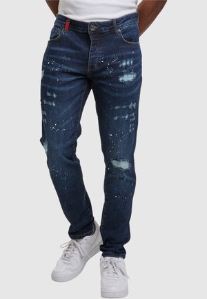 Blaue Jeans aus Denim mit einem distressed Design, Farbklecksen und Rissen in schlanker Passform. Kombiniert mit weißen Sneakers.