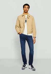 Beżowa kurtka typu bomber z zamkiem błyskawicznym, biała koszulka z nadrukiem pod spodem, ciemnoniebieskie jeansy oraz czarno-białe trampki.