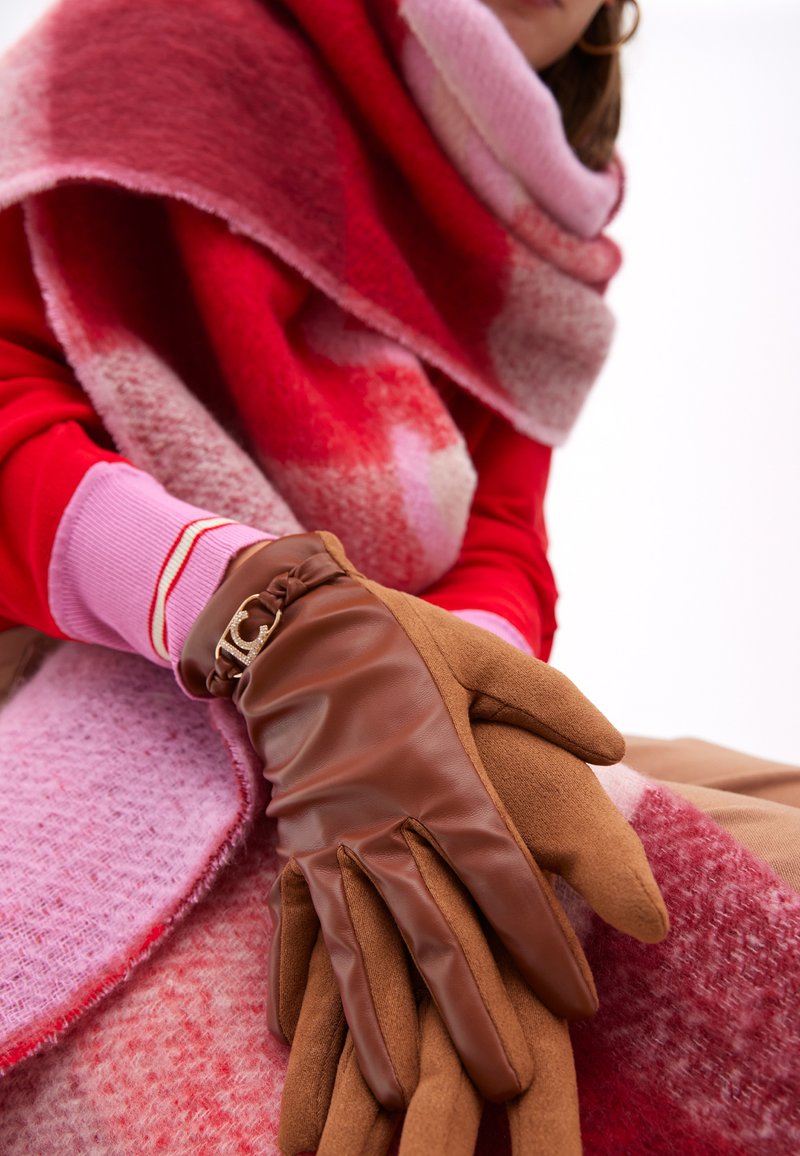 Bruna läderhandskar med ett texturerat brunt tyg vid handleden. En rosa och röd gradientscarf med fluffig textur hänger i bakgrunden.