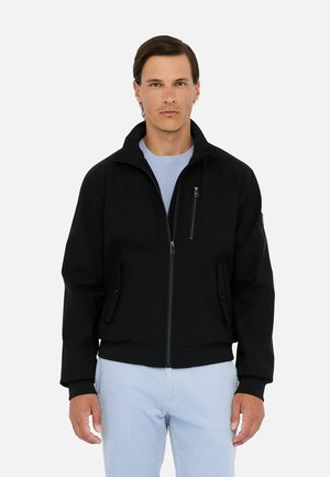 Man standing facing forward wearing a black zip-up jacket over a light blue shirt and light blue pants against plain background.