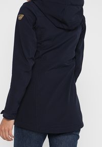 Veste softshell bleu marine avec capuche, de coupe ajustée, dotée de poches zippées et d'un logo sur l'épaule gauche.