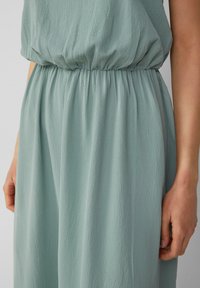 Helles grünes, strukturiertes Kleid mit einem elastischen Bund. Der Stoff hat einen leichten Glanz und einen lockeren Sitz, der sanft um die Taille und Hüften fällt.