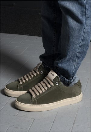 Persona con zapatillas de ante verde oliva con cordones beige y suelas blancas, de pie sobre un suelo de baldosas gris claro.