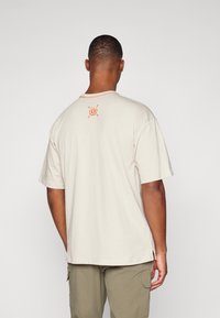 Camiseta de algodón beige con un corte relajado, mangas cortas y escote redondo. Presenta detalles en naranja y un pequeño emblema en la parte posterior.