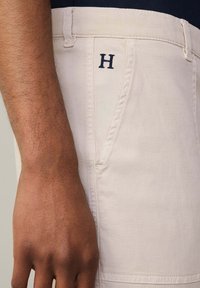 Bež pamučne kratke hlače s prednjim džepom, ukrašene plavim vezom "H" logom na pojasu. Glatka tekstura, čist dizajn.