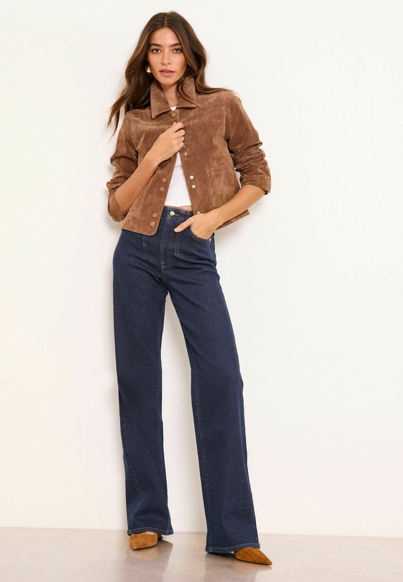 Barna szarvasbőr dzseki magas gallérral és fehér gombokkal, sötétkék magas derekú farmerrel és barna hegyes orrú cipővel párosítva. Sima textúra.