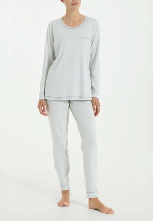 Donna che indossa una maglia a maniche lunghe grigio chiaro con tasca e pantaloni aderenti coordinati, in piedi a piedi nudi su sfondo bianco.