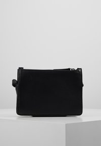 Borsa in pelle nera con chiusura a zip, posizionata su un piedistallo bianco davanti a uno sfondo semplice. Design compatto, stile minimalista.