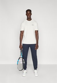 Άσπρο T-shirt με κοντά μανίκια και λογότυπο στο στήθος, συνδυασμένο με σκούρο μπλε παντελόνι τζόγερ και λευκά αθλητικά παπούτσια. Κρατάει ρακέτα τένις.