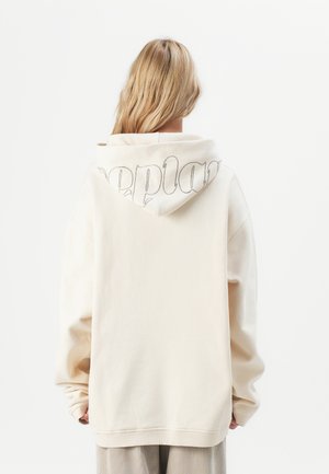 Person med blondt hår iført en overdimensjonert off-white hettegenser med delvis synlig svart tekst på hetten, stående mot en enkel hvit bakgrunn.