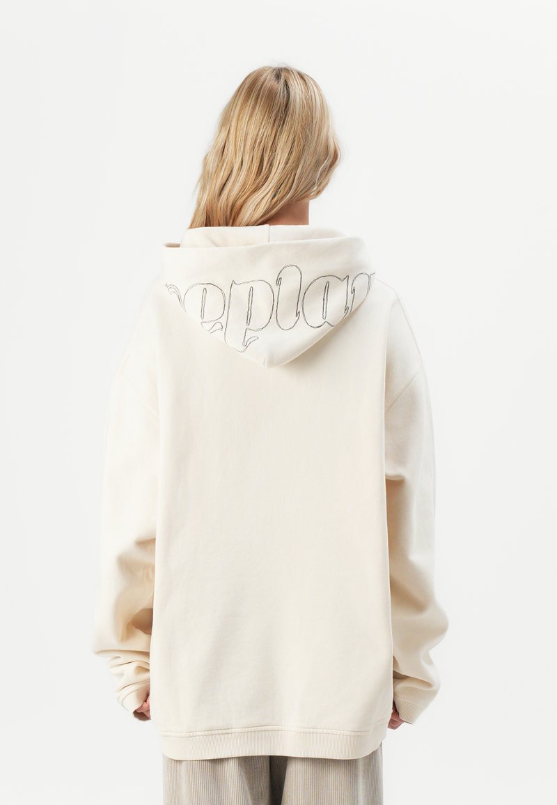 Blond persoon die een oversized off-white hoodie draagt met gedeeltelijk zichtbare zwart omlijnde tekst op de capuchon, staande tegen een effen witte achtergrond.