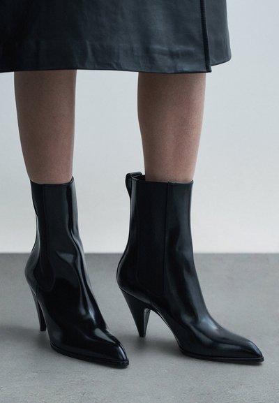Czarne skórzane botki z odsłoniętą kostką, z ostrym noskiem i wysokim obcasem typu szpilka. Posiadają elastyczne panele boczne oraz gładkie, błyszczące wykończenie.
