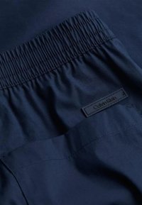 Pantaloni corti blu navy con vita elastica arricciata e un'etichetta nera con la scritta "Calvin Klein". Tessuto liscio con dettagli di cucitura minimi.