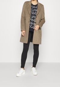 Braun karierten Mantel, navyblauen Grafikpullover mit weißem Text, schwarze Leggings und weiße Sneakers. Der Mantel hat einen Reverskragen und Knopfdetails.