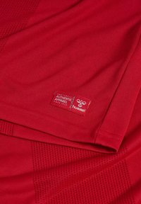 Tessuto sportivo rosso con pannelli testurizzati e una piccola etichetta rossa con la scritta "Authentic Apparel" e il logo "hummel".