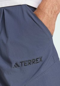 Pantalones cortos deportivos azules con una textura suave, que cuentan con un bolsillo lateral y un logotipo bordado en negro "TERREX". El diseño es funcional y minimalista.