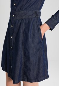 Tummansininen farkku mekko, jossa on napitettu etuosa, pitkät hihat, kapea vyötärö ja sivutaskut. Sileä, kevyt rakenne.