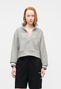 Νεαρό άτομο με κοντά κόκκινα μαλλιά, που φοράει ανοιχτογκρί φούτερ με φερμουάρ και μαύρα σορτς Calvin Klein, στέκεται μπροστά σε λευκό φόντο.