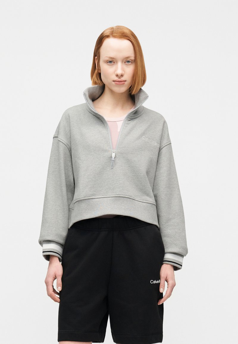 Νεαρό άτομο με κοντά κόκκινα μαλλιά, που φοράει ανοιχτογκρί φούτερ με φερμουάρ και μαύρα σορτς Calvin Klein, στέκεται μπροστά σε λευκό φόντο.