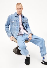 Lichtblauwe denim jas met twee borstzakken, gecombineerd met een roze en grijze geruite shirt, en losse lichtblauwe jeans. Zwarte schoenen en gestreepte sokken.