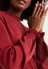 Haut en maille rouge avec un col montant, des manches bouffantes et des poignets froncés. Les poignets sont ornés de petits boutons décoratifs dorés. Texture douce avec un léger éclat.