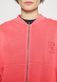Sudadera con cremallera en color coral, fabricada en algodón y con cuello de canalé. Presenta un detalle de logo bordado y una cremallera metálica en la parte delantera.
