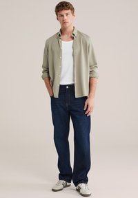 Hellgrünes Hemd über einem weißen Tanktop, kombiniert mit dunkelblauen Jeans und weißen Turnschuhen mit grünen Streifen. Lässige Passform und entspannter Stil.