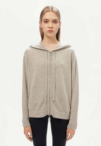 Ljusgrå zip-hoodie i mjukt tyg, med dragsko på huven, långa ärmar och en avslappnad passform. Förstärkt fåll och muddar.