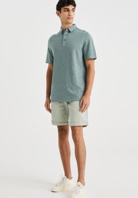 Polo shirt albastru deschis, texturat, cu guler și trei butoane, asociat cu pantaloni scurți verzi deschis și adidași albi.