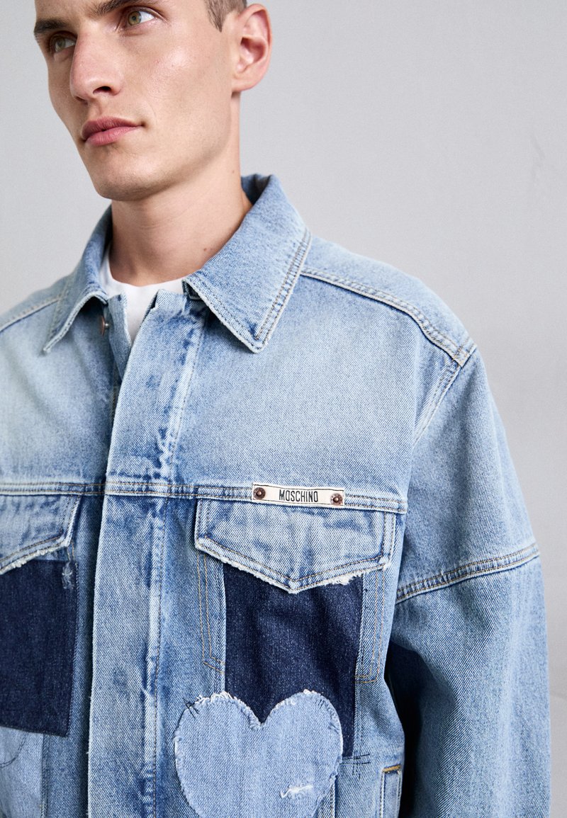 Jongeman draagt een lichtblauwe denimjack met donkerblauwe zakken, een hartvormige patch op de onderste zak en een Moschino-label op de borst.