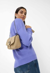 Helllila gestreifter Pullover und beige Wildleder-Handtasche mit einer Ketten-Umhängegurt, seitlich gezeigt. Dunkle Jeans vervollständigen das Outfit.