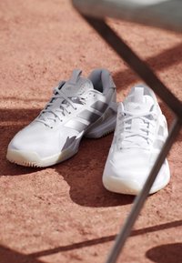 Witte Adidas sportschoenen met zilveren strepen geplaatst op roodachtige buitengrond onder beschaduwde metalen staven.