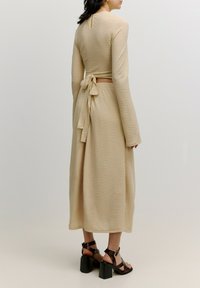 Beige langärmliges Kleid aus strukturiertem Stoff, mit einem Rückenschleifen-Detail und einem Schlüssel-Ausschnitt. Kombiniert mit schwarzen hohen Sandalen.