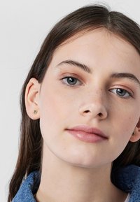 Pendientes de oro en forma de oso con acentos verdes. El modelo tiene piel clara y cabello largo y castaño, llevando una chaqueta de mezclilla.