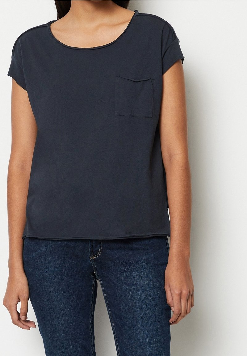 Donna che indossa una camicia a maniche corte di un blu navy scuro con taschino e jeans blu scuro, in piedi contro uno sfondo bianco semplice.