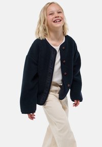 Veste en polaire bleu foncé avec une texture douce, de grandes manches et des boutons marron. Conçue avec un col rond et finie avec une bordure contrastante.