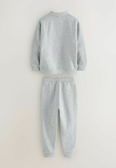 Pilkas džemperis ir treniruotės kelnės, pagamintos iš minkštos medžiagos. Džemperis turi aukštą apykaklę; kelnės turi elastingą juosmenį ir rankogalius.
