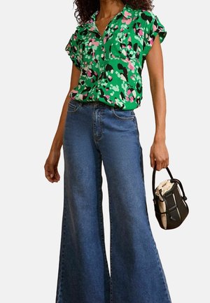 Kvinde iført en grøn bluse med korte ærmer og blomsterprint, højtaljede, vidde blå jeans og med en sort- og beige håndtaske.