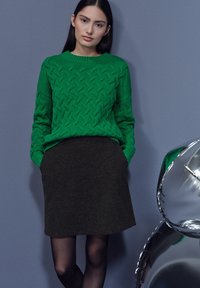 Groen gebreide trui met een gestructureerd patroon, gecombineerd met een donkere rok. Grijze achtergrond, gedeeltelijk zichtbare reflecterende metalen accenten.