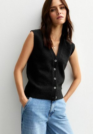 Cardigan nero senza maniche con scollatura a V, chiusura con bottoni e una texture morbida, abbinato a jeans azzurri a vita alta.