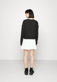 Melnā apgrieztais pel�ēkā krekls ar nokritušām plecu daļām, saskaņots ar baltu krokotu svārku, uzvilkts ar melnām zeķēm un kedām, skatoties no mugurpuses.