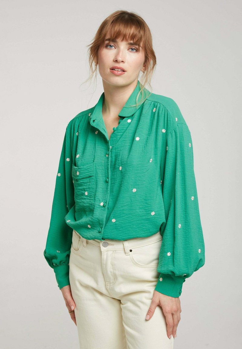 Chemisier vert à boutons, en tissu léger, avec des broderies discrètes de fleurs blanches, coupe décontractée et manches ballon.