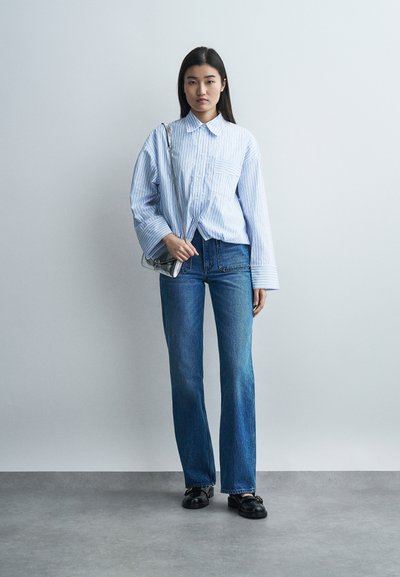 Νεαρή γυναίκα στέκεται μπροστά σε έναν μονόχρωμο τοίχο, φορώντας μπλε ριγέ φαρδιά πουκάμισο, μπλε τζιν, μαύρα παπούτσια και κρατώντας μια ασημένια τσάντα.